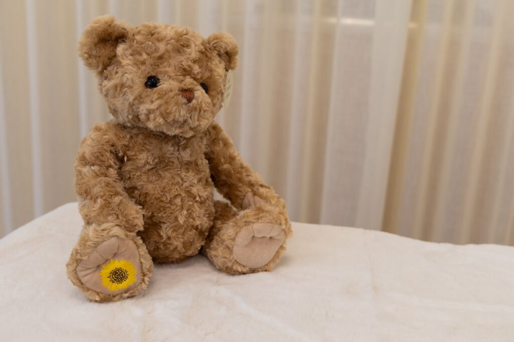 Beige furry teddy bear sitting up on white sheets, with a flower logo on the left foot.