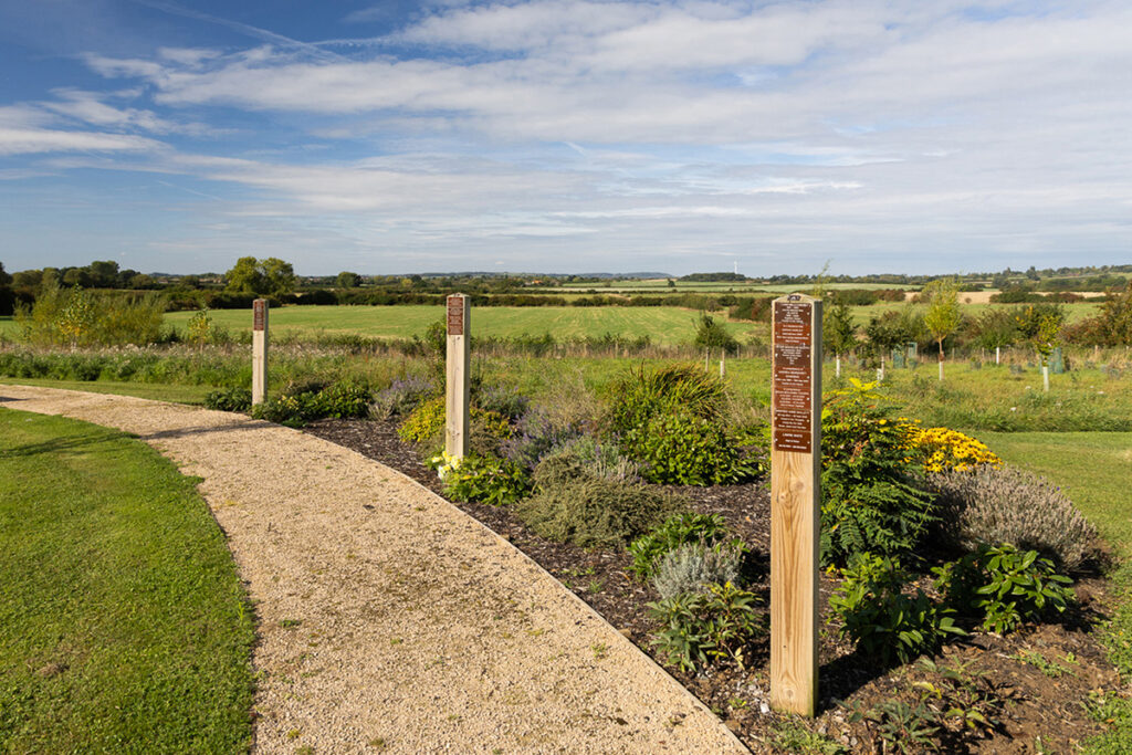 Bierton memorial garden. Wooden memorial markers along a footpath. On the right, bushes and grass surround behind.